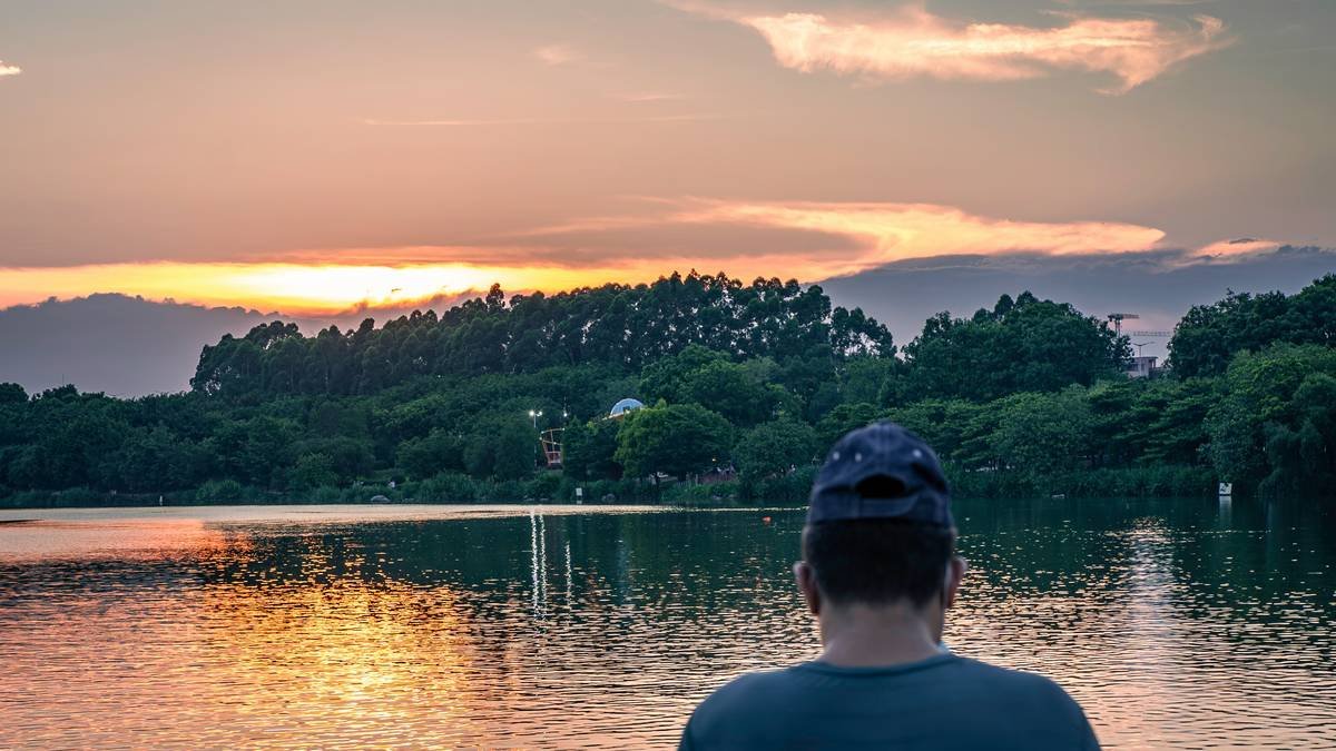 From behind, a person wearing a cap gazes out at a serene lake during sunset. The calm water reflects the soft, warm colors of the sky, creating a peaceful and contemplative scene perfect for illustrating themes of solitude, reflection, and finding tranquility in nature.